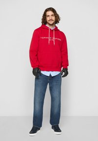 Felpa rossa con logo bianco, tasca frontale e lacci. Indossata sopra una maglietta azzurra, abbinata a jeans blu e scarpe da ginnastica scure.