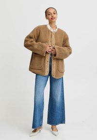 Brun fleecejacka med stora fickor, knappstängning och en strukturerad yta, parat med vida blå jeans och vita klackar.