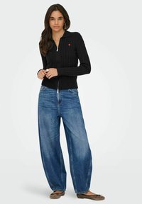 Kobieta w czarnym swetrze zapinanym na zamek z warkoczem, z małym czerwonym sercem, w szerokich niebieskich dżinsach i beżowych balerinach, stojąca na białym tle.