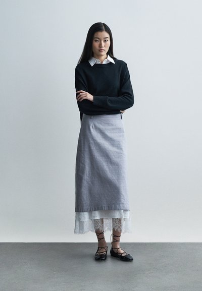 Pull noir en tricot, jupe en lin grise, ourlet en dentelle blanche, ballerines noires à brides croisées. Le mannequin est debout, les bras croisés, sur un fond neutre.