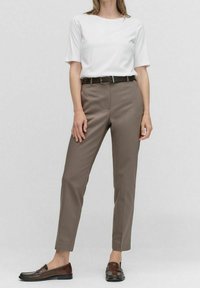 Kobieta w białej koszuli z krótkim rękawem, taupe prostych spodniach z czarnym paskiem oraz ciemnobrązowych skórzanych mokasynach, stojąca na jednolitym tle.