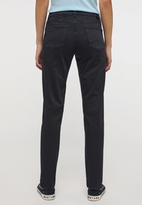 A modell hátulról látható, sötét farmernadrágot és "MUSTANG" márkájú fekete sportcipőt visel; a háttér sima és egyszerű.