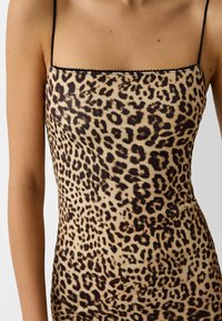 Camisole s leopardím vzorem vyrobená z hladkého materiálu. Má tenké černé ramínka a šikmý výstřih. Béžový základ s černými skvrnami.