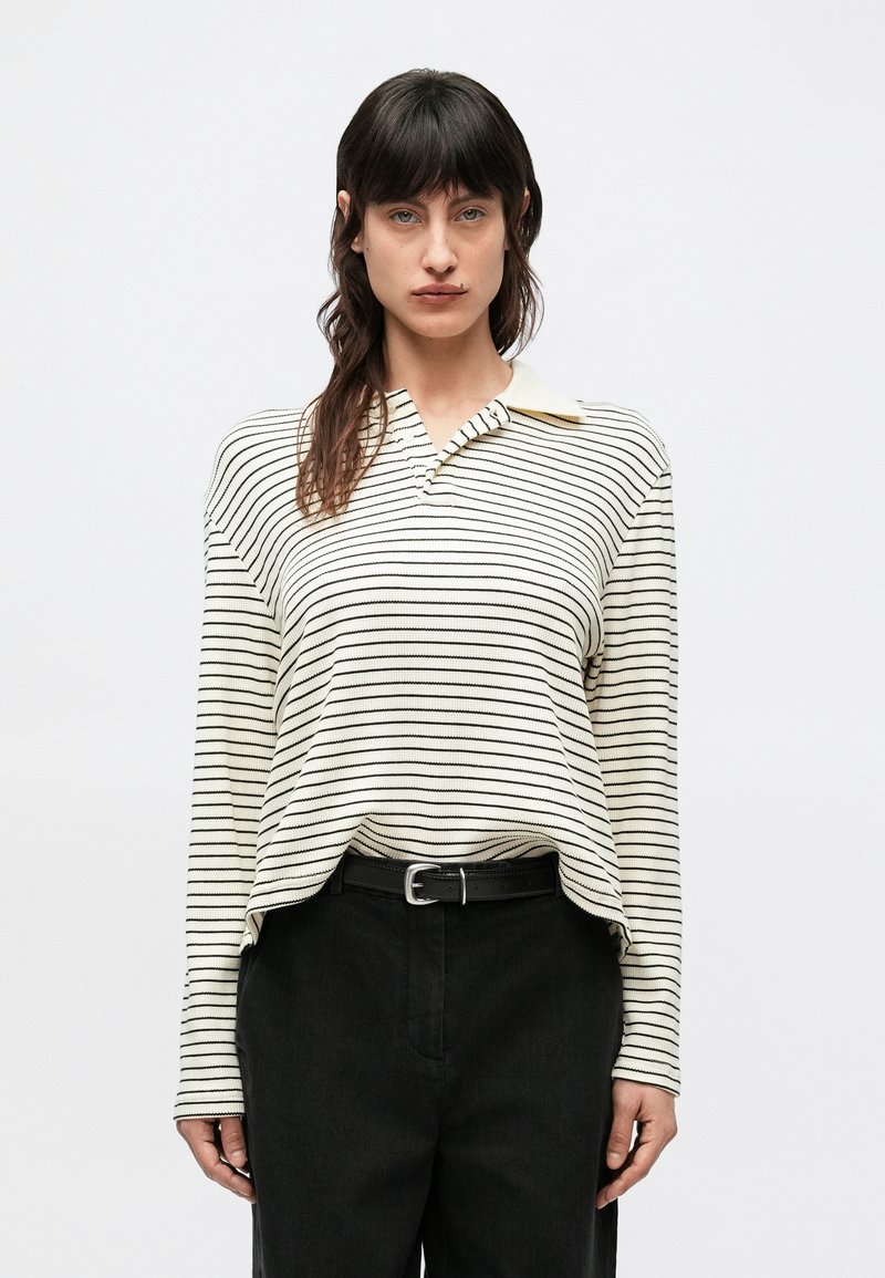 Dlhý rukáv, blúzka s golierom v krémovej farbe s čiernymi pruhmi. Má voľný strih a textúrovanú látku. Kombinovaná s čiernymi nohavicami s vysokým pásom.