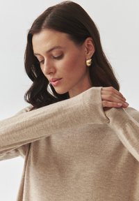 Beige gebreide trui met lange mouwen en geribde manchetten, gedragen met gouden getextureerde oorbellen, model kijkt naar beneden tegen een lichte achtergrond.