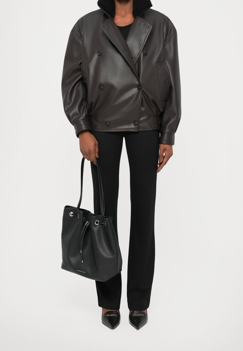 Personne portant une veste en cuir marron foncé, un pantalon noir et des chaussures noires pointues, tenant un sac à main en cuir noir par la poignée.