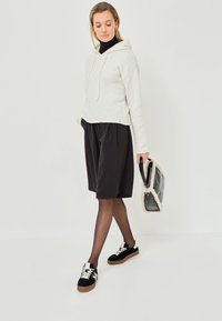 Vit, texturerad huvtröja med dragsko, i kombination med svarta, veckade shorts och svarta sneakers med cremefärgade detaljer. Bär en fluffig, svart handväska.