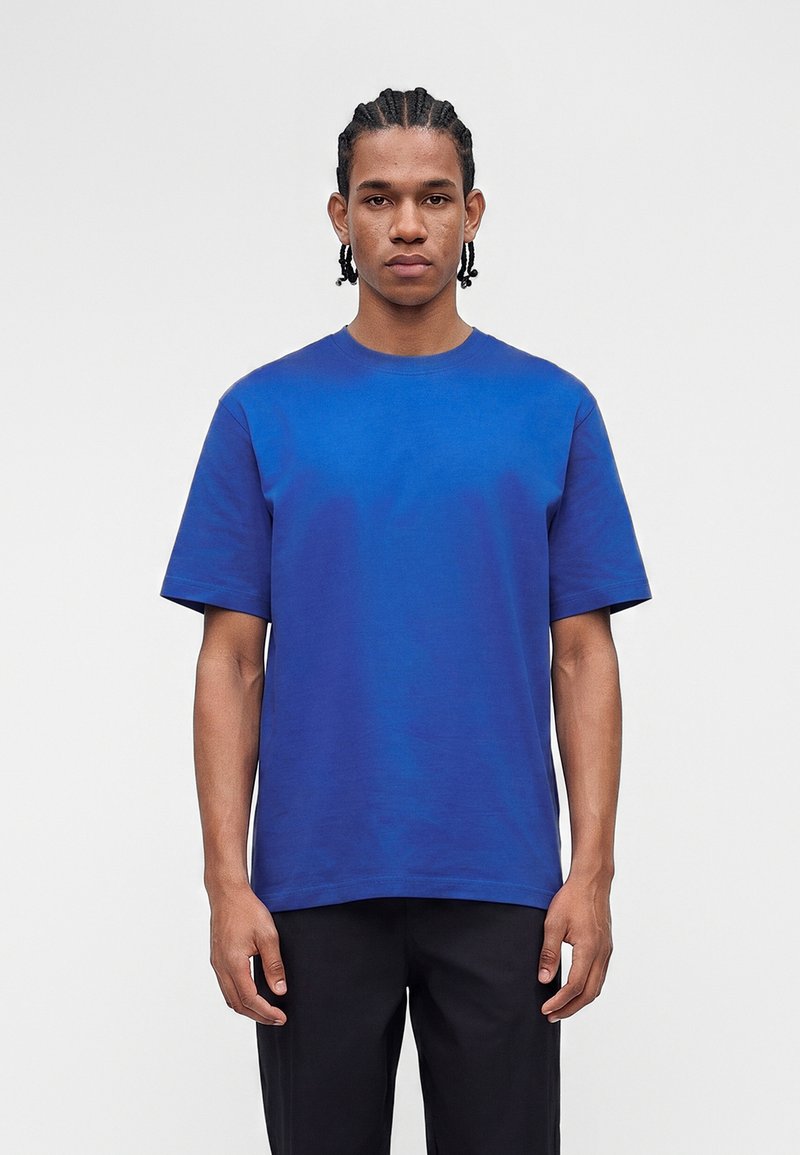 Νέος άνδρας με πλεξούδες, ντυμένος με απλό μπλε κοντομάνικο μπλουζάκι και μαύρο παντελόνι, στέκεται μπροστά σε απλό ανοιχτό φόντο.