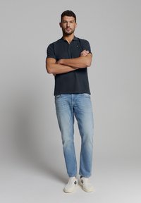 Donkerblauwe polo, lichtblauwe spijkerbroek, witte sneakers. Model staat met armen gekruist tegen een effen achtergrond. Gepolijste uitstraling, casual stijl.
