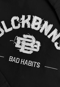 Zwarte stoffen patch met witte geborduurde tekst "BLCKBNS" en "BAD HABITS," met een versleten textuur en prominente letters.
