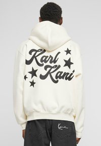 Kremfarget hettegenser med store sorte "Karl Kani" bokstaver og svarte stjerner på baksiden; myk tekstur, avslappet passform.