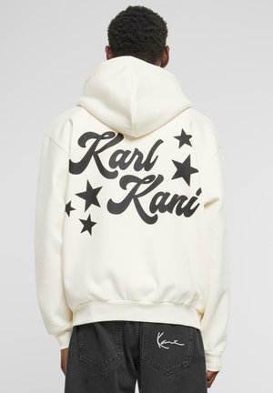 Sweatshirt à capuche crème avec de grandes lettres noires "Karl Kani" et des graphiques d'étoiles noires au dos ; texture douce, coupe décontractée.