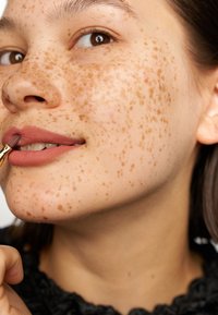 Zbliżenie na twarz z jasnobrązowymi piegami, aplikującą kolor do ust pędzelkiem. Cechuje się miękką, gładką skórą i subtelnym matowym wykończeniem ust.