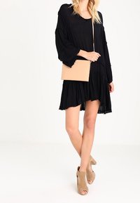 Žena v volných černých šatech drží béžovou křížem přes rameno umístěnou kabelku, má na sobě béžové sandály na odkryté špičce a stojí na bílém pozadí.