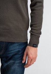 Nahaufnahme eines Oberkörpers einer Person, die einen strukturierten braunen Pullover mit einem schwarzen Etikett am Ärmel und blauen Jeans trägt.