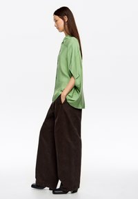 Top de satén verde con un corte holgado y aberturas laterales, combinado con pantalones anchos de algodón marrón y zapatos de tacón negros.
