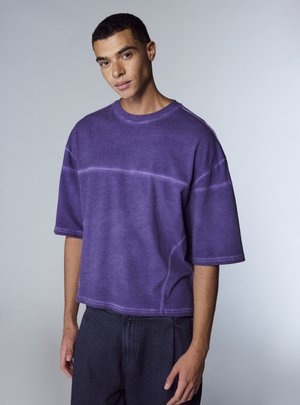 Maglietta oversize viola realizzata in tessuto morbido, con maniche corte, scollo rotondo e dettagli di cuciture distintive.