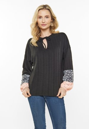 Top nero con un motivo testurizzato, caratterizzato da un design con nodo al collo. Maniche lunghe con motivi floreali in rosa e blu navy, abbinato a jeans blu.