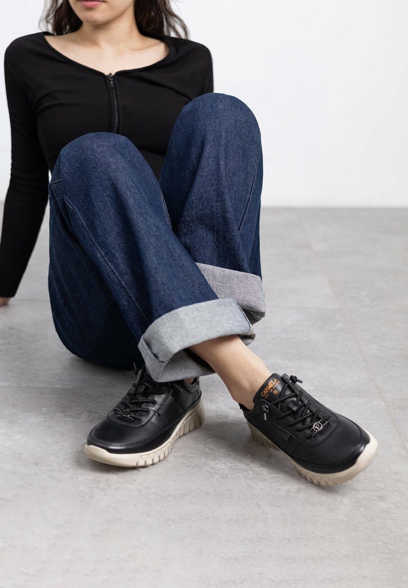 Zapatillas negras con cordones, con una parte superior texturizada y suela color beige claro, combinadas con unos vaqueros oscuros de mezclilla remangados que tienen un puño gris claro.