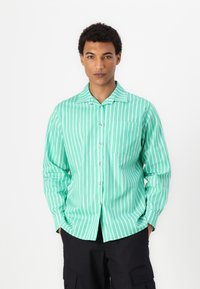 Camisa de botão verde-claro com riscas brancas verticais, colarinho pontiagudo, mangas longas e um bolso no peito. Textura de tecido suave.