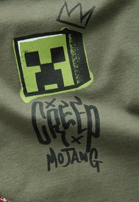 Olivegrünes Baumwoll-T-Shirt mit einem großen pixelierten grünen Creeper-Gesicht-Logo und schwarzem Graffiti-Text mit einer Krone-Illustration.