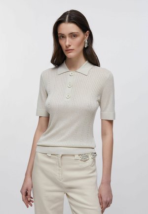 Donna che indossa una polo a maniche corte beige a coste con tre bottoni e pantaloni chiari decorati con strass sulla tasca.