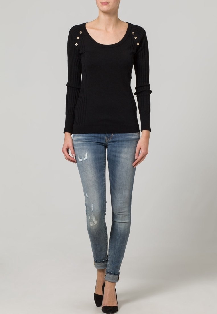 Top noir à motifs côtelés à manches longues avec un col rond et des accents de boutons sur les épaules, associé à un jean skinny déchiré bleu clair et des talons noirs.