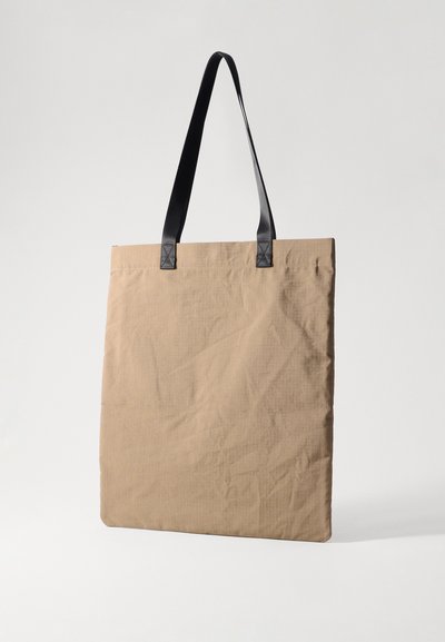 Tote bag made of textured beige fabric with black flat handles. Simple rectangular design, featuring minimal stitching details.