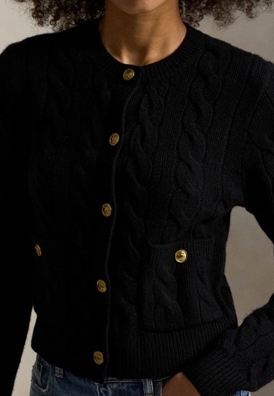 Kvinde iført en sort kabelstrikket cardigan med guldknapper og en lomme foran, kombineret med blå denimjeans.
