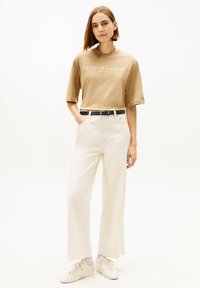 Beige T-Shirt mit kurzen Ärmeln und weißem Logo, hoch taillierte cremefarbene Hose mit weitem Bein, schwarzer Gürtel und weiße Sneakers mit strukturierten Sohlen.