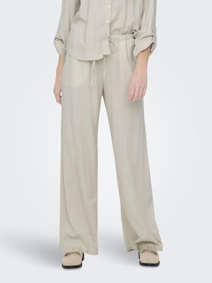 Femme portant un pantalon beige à jambes larges et une chemise à manches longues assortie retroussée, associée à des mocassins beige sur fond blanc.
