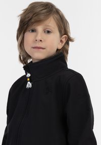 Svart zip-up jacka med hög krage, broderad logotyp och dekorativt fransdetalj. Tillverkad av mjukt, lätt tyg.