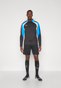 Svart sports-langermet topp med blå og hvite detaljer, kombinert med svarte shorts, svarte knestrømper og svarte sko med hvit logo.