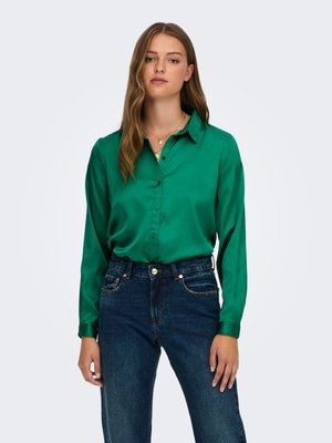 Jeune femme aux longs cheveux châtain clair porte un chemisier vert brillant boutonné et un jean bleu foncé taille haute sur un fond uni.
