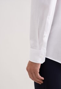 Biała koszula z zapinanym na guziki mankietem, gładką fakturą i dopasowanym krojem, zbliżenie na rękaw i dłoń.