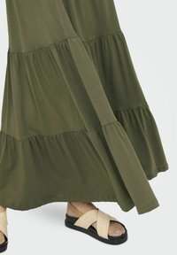 Vestido maxi de color verde oliva con capas y tejido texturizado, combinado con sandalias negras con tiras beige cruzadas y suela plana.