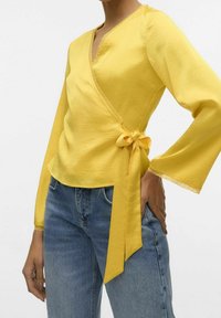 Žlutá blůza s dlouhými rukávy, krajkovým detailem a stahováním v pase, spárovaná s modrými džíny. Jemná, hladká textura látky.