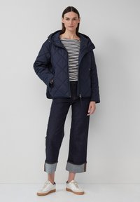 Marineblauwe gewatteerde jas met capuchon, voorzien van voorzipper en zijzakken. Gedragen over een gestreept shirt en gecombineerd met donkerdenim en witte sneakers.