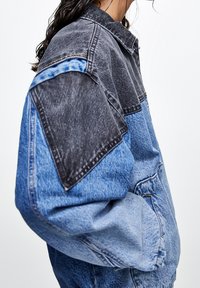 Chaqueta vaquera con diseño bicolor, con hombros gris oscuro y cuerpo, costuras de los hombros y bolsillos en azul claro. Tejido texturizado.