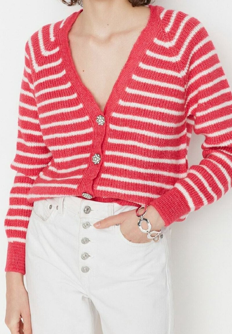 Femme portant un cardigan à rayures rouges et blanches avec des boutons floraux, associé à un jean blanc taille haute et un bracelet en maillons argentés.