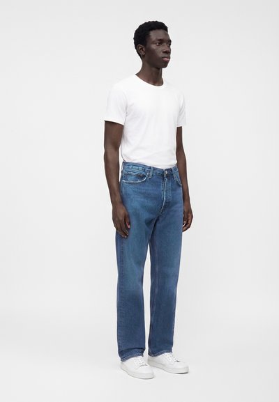 Homme debout, légèrement tourné vers la droite, portant un t-shirt blanc, un jean bleu et des baskets blanches sur un fond blanc uni.
