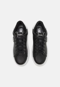 Schwarze Ledersneaker mit runder Spitze, flacher weißer Gummisohle, schwarzen Schnürsenkeln und einem silbernen Akzent an der Ferse. Branding auf der Zunge sichtbar.