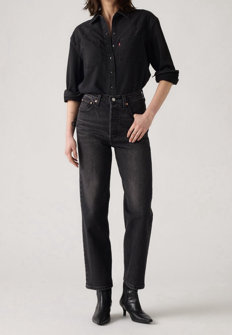 Femme portant une chemise en jean noire aux manches retroussées, un jean noir taille haute et des bottines à talons noires, debout devant un fond uni.