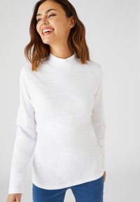 Witte langemouwen top met een hoge hals, gemaakt van zacht katoen. Kenmerkt zich door een gladde textuur en een ontspannen pasvorm, gedragen met blauwe jeans.
