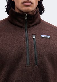 Κοντινό πλάνο ενός ατόμου που φορά καφέ fleece Patagonia με ψηλό γιακά και δύο κάθετες μαύρες τσέπες με φερμουάρ στο στήθος.