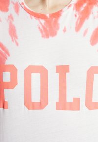 Biała koszulka z wzorem tie-dye w kolorze koralowym oraz wyrazistym napisem "POLO" w koralowym kolorze. Materiał wygląda na miękki, z klasycznym okrągłym dekoltem.