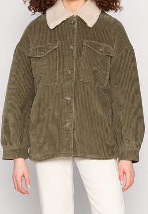 Vrouw draagt een olijfgroene corduroy jas met een geïmplementeerde kraag van schapenvacht en knoopsluiting, gecombineerd met off-white broek.