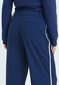 Námornické modré široké kalhoty s hladkou texturou, s bílými bočními pruhy a zadní kapsou. Vydané s pleteným modrým svetrem.