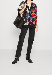 Strickjacke mit mehrfarbigen Herz- und Blumenmustern auf schwarzem Grund, kombiniert mit schwarzen Jeans und schwarzen Stiefeln mit Absatz. Schwarze Handtasche enthalten.