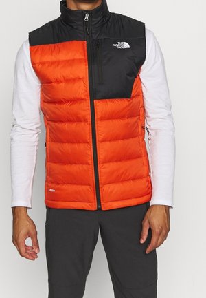 Mann trägt eine orange-schwarze gepolsterte ärmellose Weste über einem weißen Langarmhemd und dunkler Hose, steht vor einfachem Hintergrund.